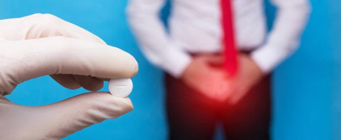 Arzt hält Medikament in die Kamera - im Hintergrund ein Mann mit Schmerzen im Genitalbereich