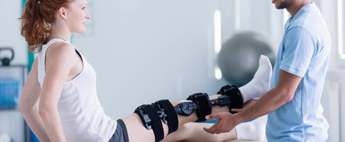 Physiotherapeut behandelt Frau mit Knieschiene auf Liege
