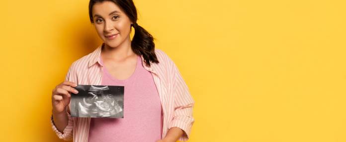 Schwangere Frau fasst sich mit einer Hand an den Bauch - in der anderen Hand hält sie ein Ultraschallbild