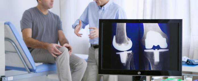 Mann im Gespräch mit Orthopäden - im Vordergrund Röntgenaufnahme am Computer mit Knieprothese