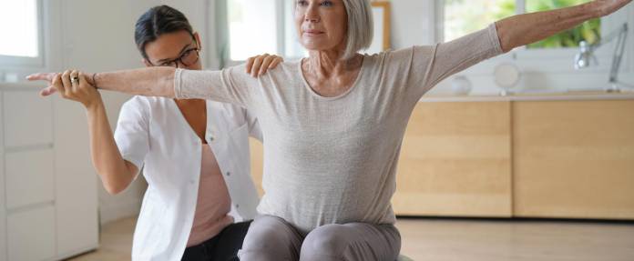 Seniorin bei Übung auf dem Gymnastikball unter Anleitung einer Therapeutin