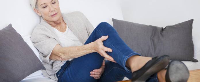 Ältere Frau, die auf dem Sofa sitzt, mit Schmerzen in den Beinen