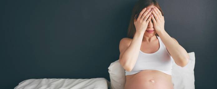 Schwangere Frau sitzend im Bett schlägt Hände vor die Augen