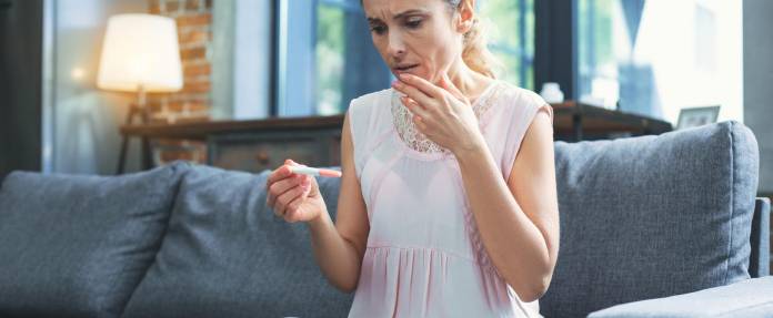 Reifere Frau sitzt geschockt auf dem Sofa mit einem Schwangerschaftstest in der Hand